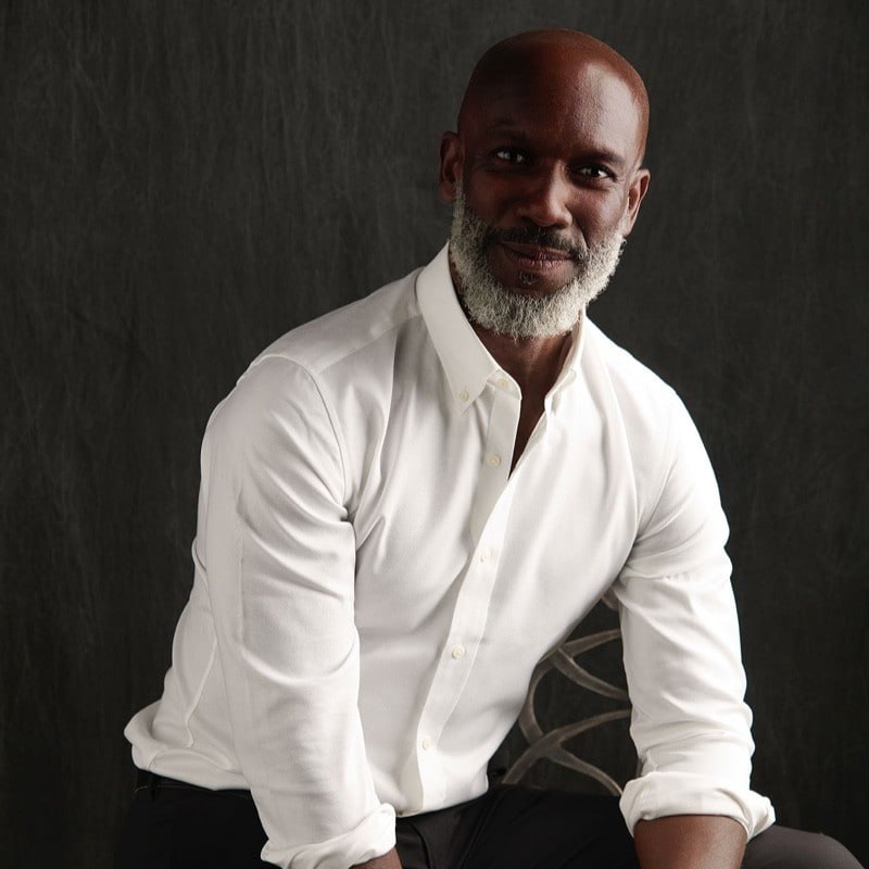 Rodney Hutton, seated in a white shirt against a charcoal studio backdrop.