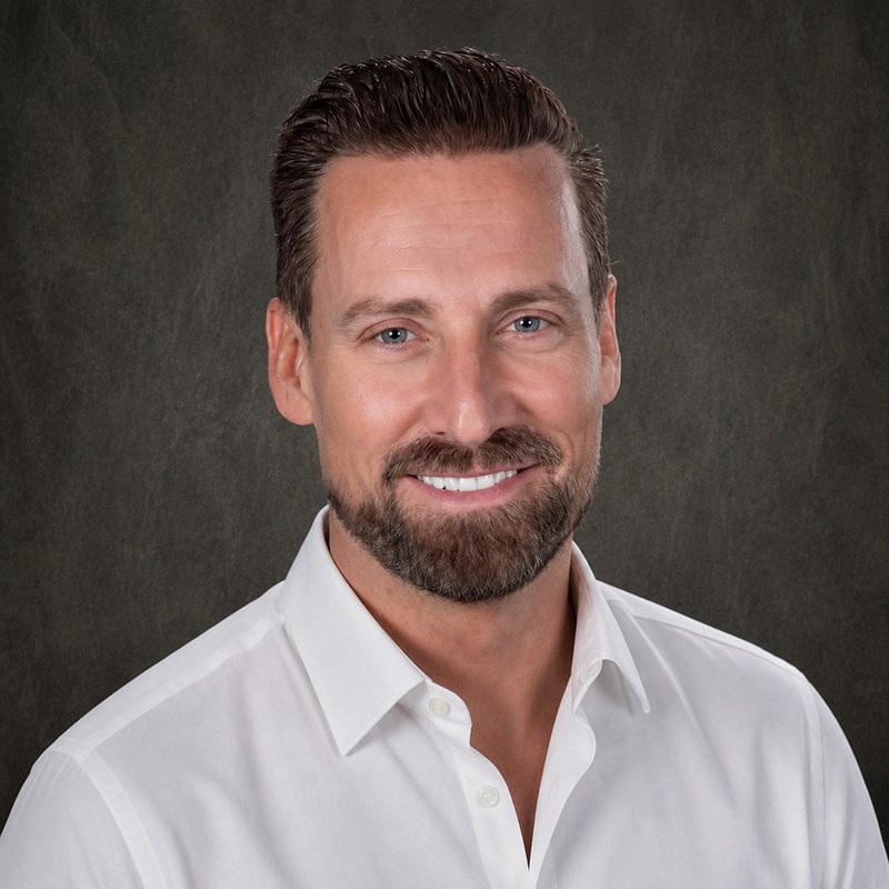 Sam Bremner, in a white shirt against a charcoal studio backdrop.